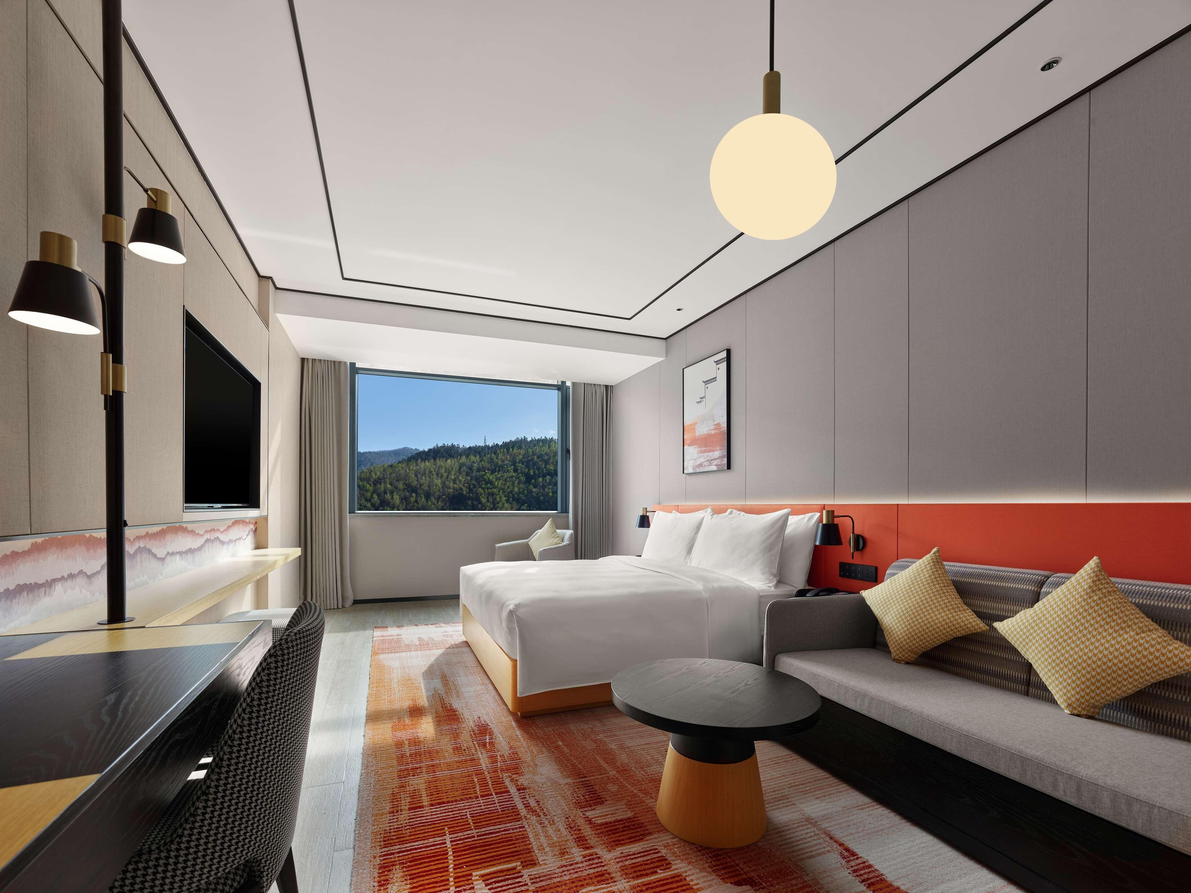 Une chambre d’hôtel moderne dotée d’une grande fenêtre, d’un lit, d’un canapé, d’une petite table et d’un téléviseur.