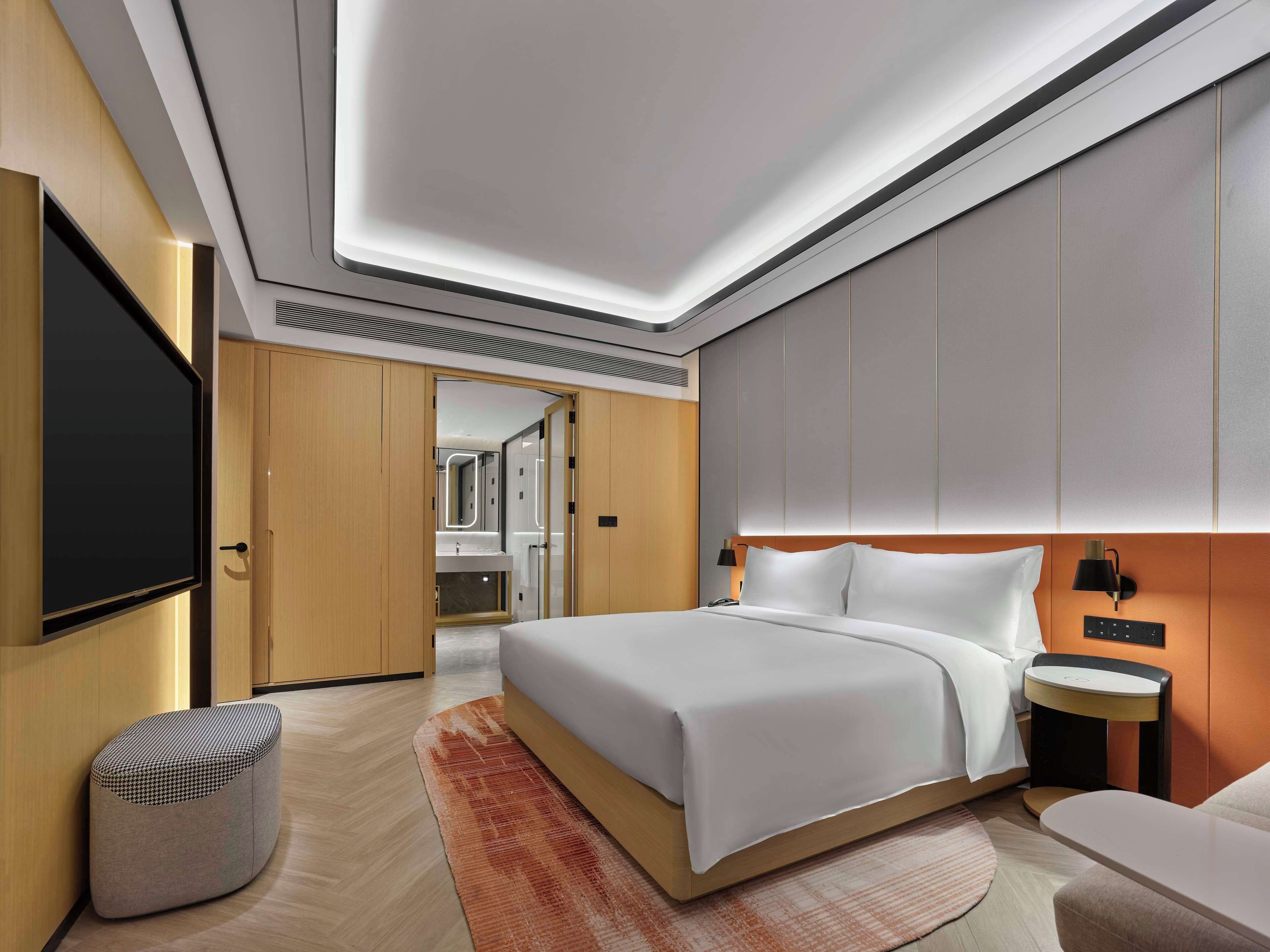 A modern hotel room with a large bed, a bedside table, a TV, and a bathroom visible through an open door.