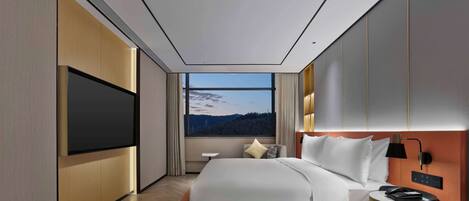 Una habitación de hotel moderna con una cama grande, un televisor de pantalla plana, una mesita de noche con lámpara y vistas a un bosque a través de un amplio ventanal.