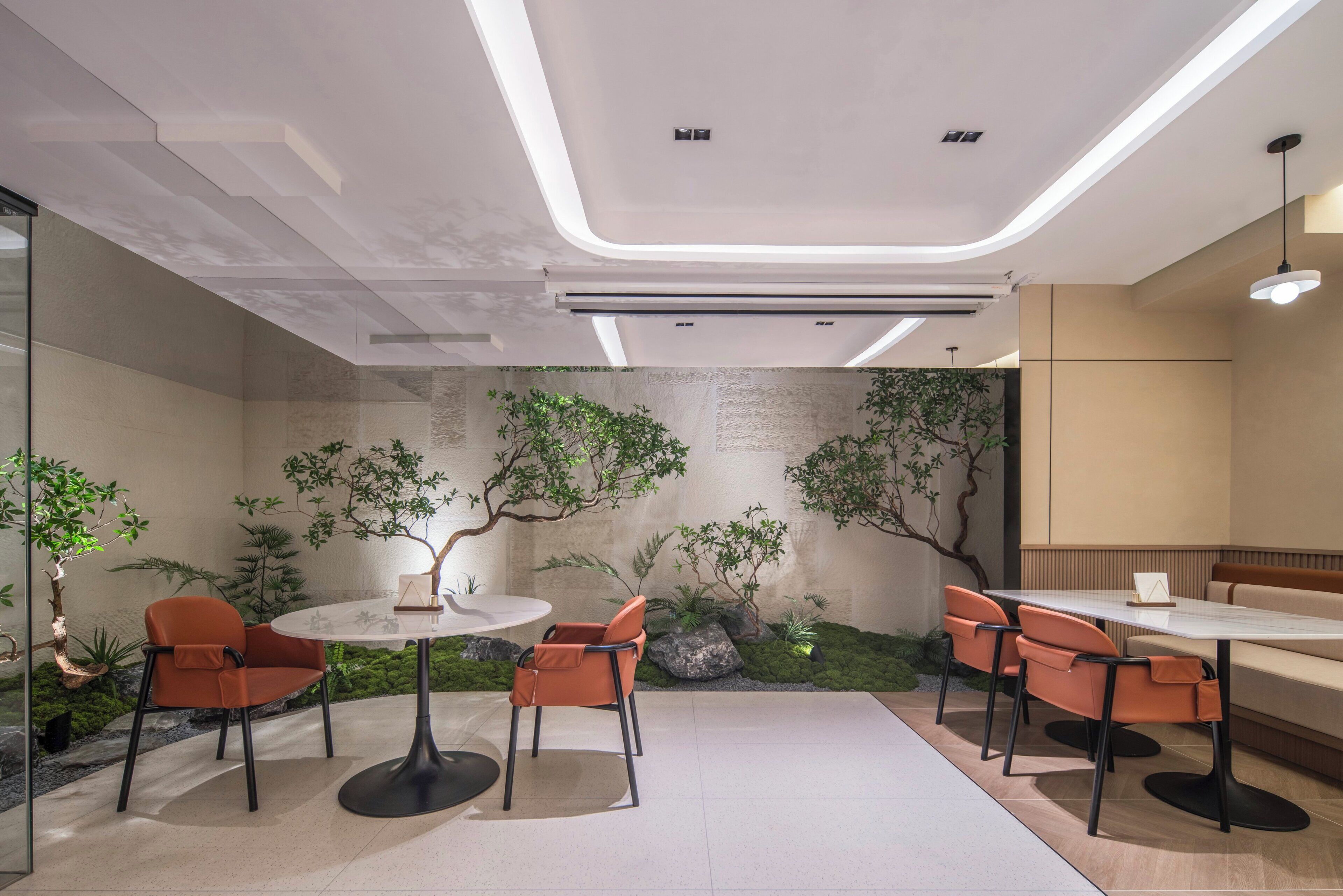 Un espacio interior moderno con una mesa redonda, cuatro sillas, un sofá y una pequeña área ajardinada con plantas.