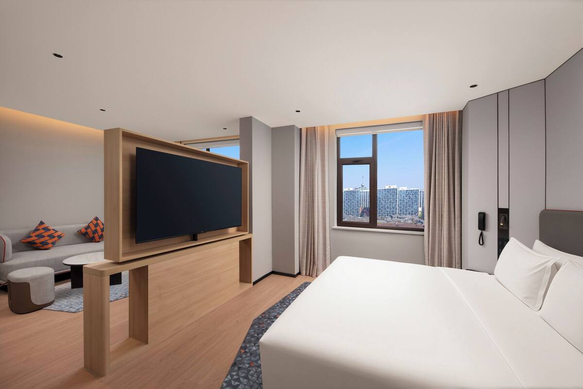 A modern hotel room with a large bed, a flat-screen TV, a sofa, and a city view.