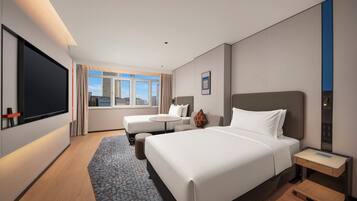 A hotel room with a large bed, a smaller sofa, a flat-screen TV, a desk, and a window with a city view.