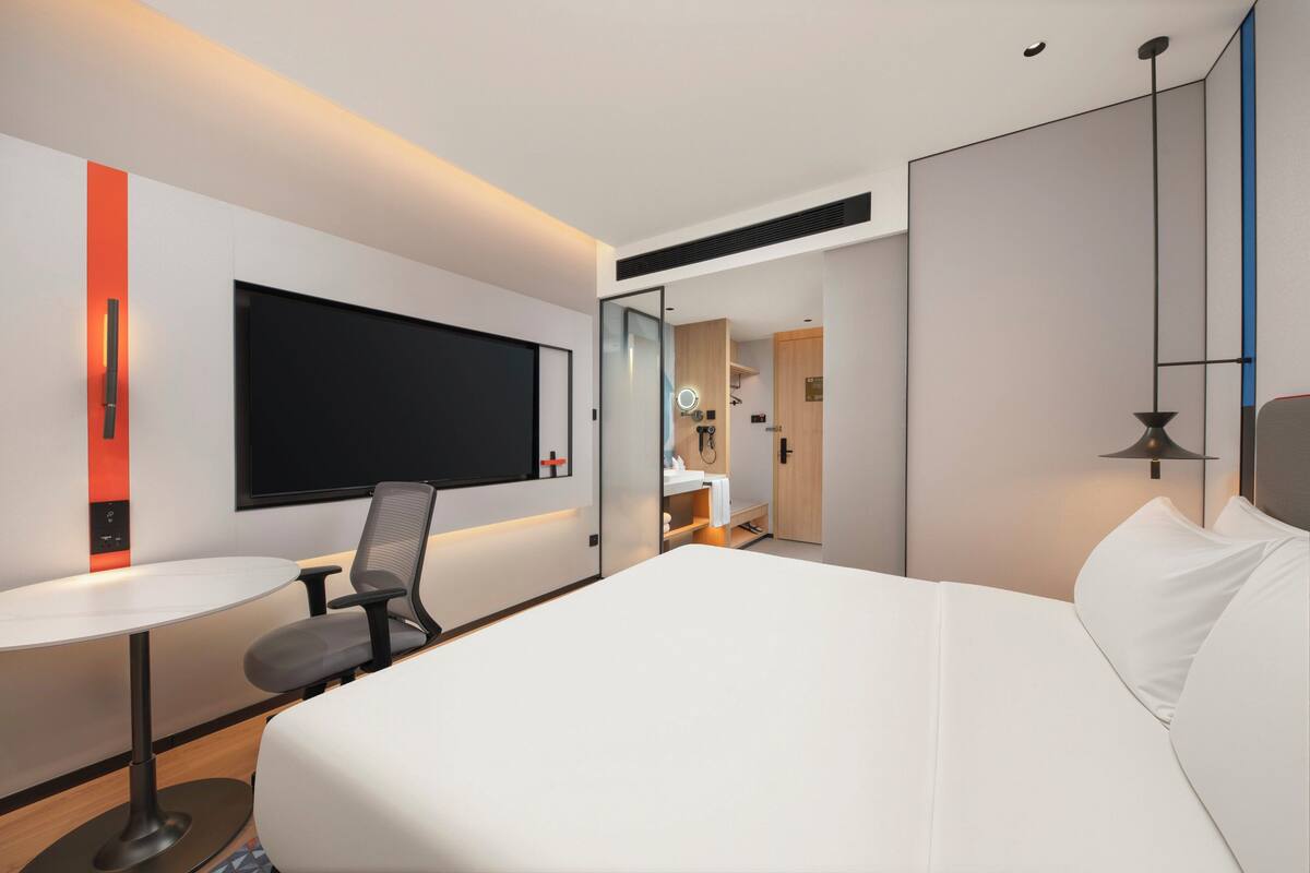 A modern hotel room with a large bed, a desk with a chair, a television, and a bathroom visible through an open door.