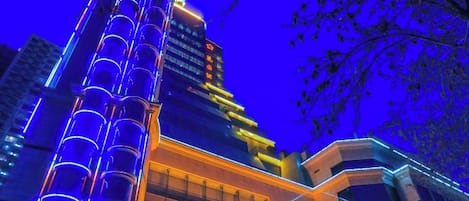 A multi-story building with a prominent vertical structure on the left and a brightly lit entrance on the right.