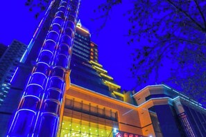 A multi-story building with a prominent vertical structure on the left and a brightly lit entrance on the right. - Korla Pear City Garden Hotel (Korla)