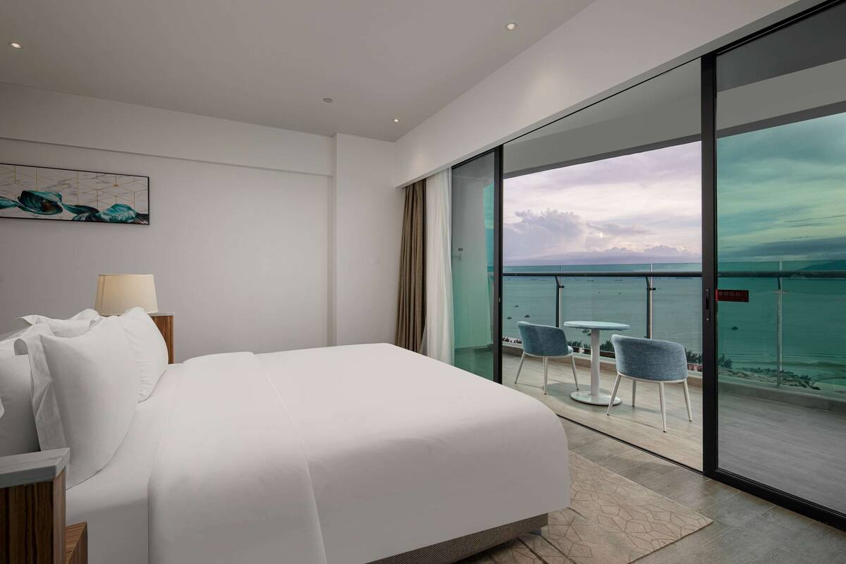 A modern hotel room with a large bed, a glass door leading to a balcony with a view of the sea and a cityscape.