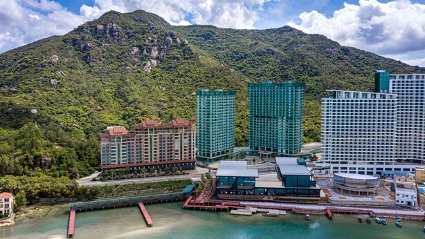 Un complejo hotelero costero con varios edificios altos, un muelle y montañas al fondo.