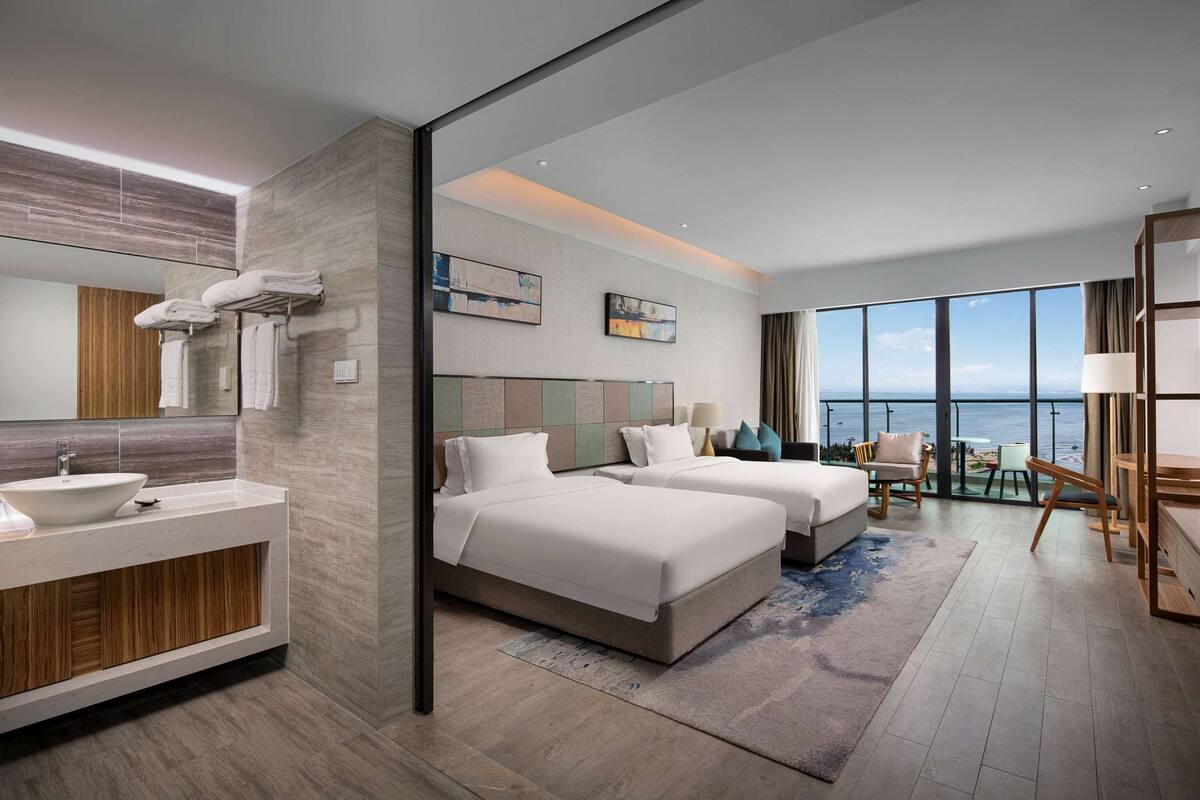 A modern hotel room with a large window, a private balcony, a large bed, a sofa, a desk, and a bathroom with a sink and mirror.