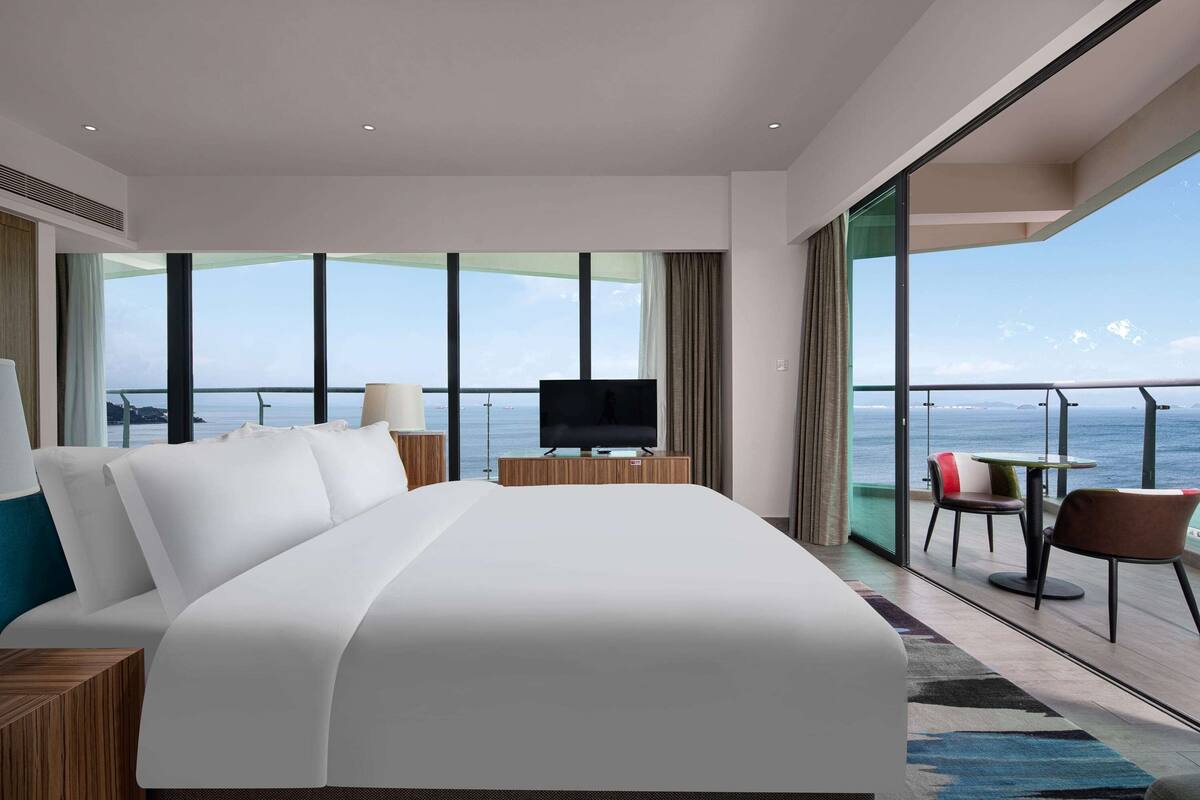 A modern hotel room with a large bed, a TV, a balcony with a table and chairs, and a view of the sea.