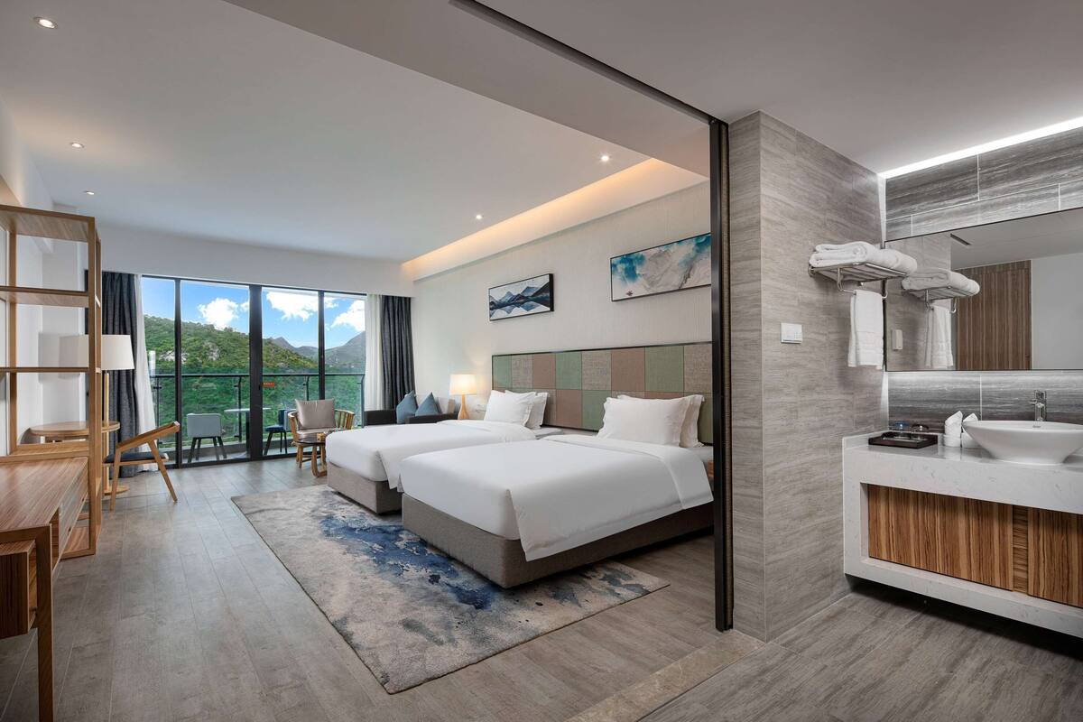 A modern hotel room with two beds, a large window with a mountain view, a balcony, a wooden desk, and a bathroom with a large mirror and sink.