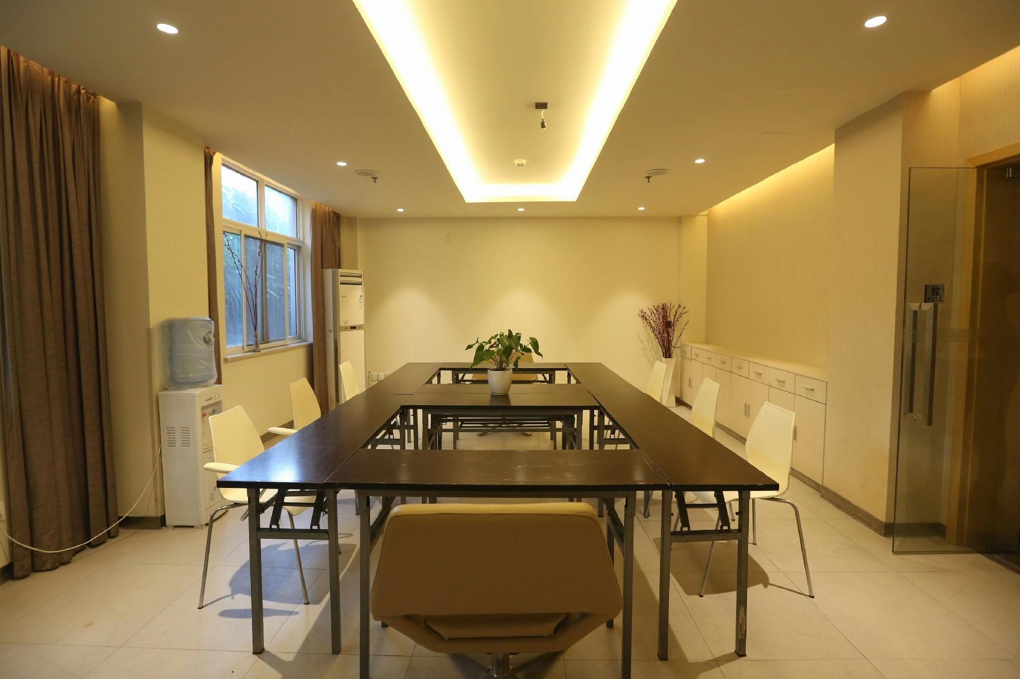 A conference room with a long table, chairs, a potted plant, a water dispenser, and a refrigerator.