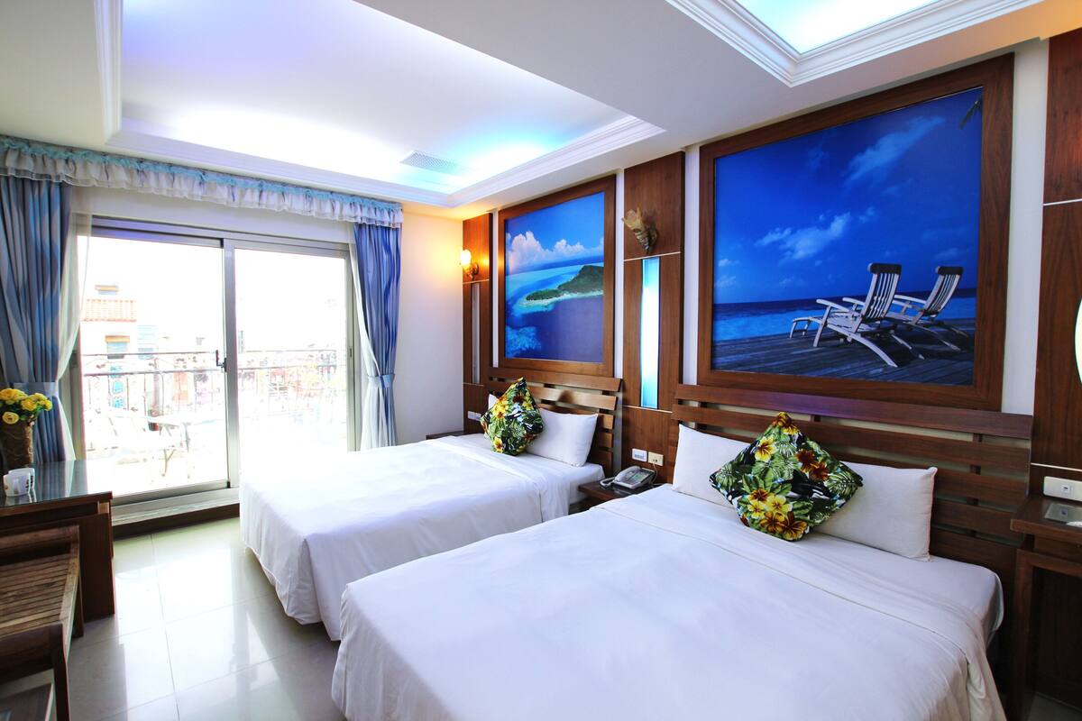 A hotel room with two beds, a balcony with a view, and a large painting of a beach scene.