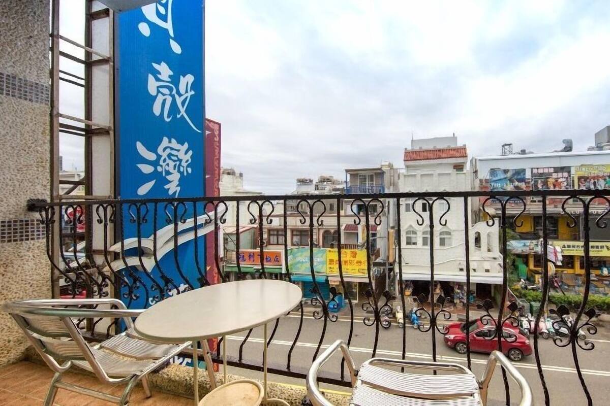 A balcony with metal chairs and a table, overlooking a busy street with shops and vehicles.