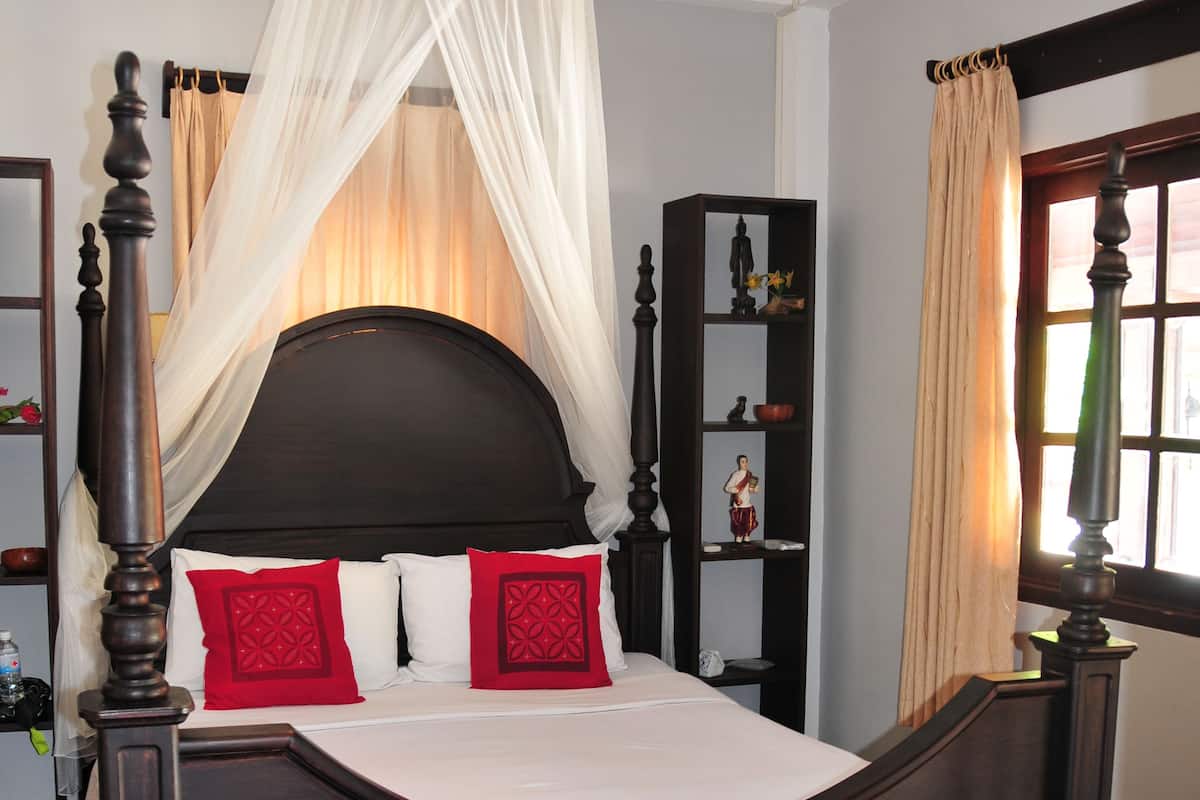 A dark wood four-poster bed with a canopy, red pillows, and a nightstand with a bottle.