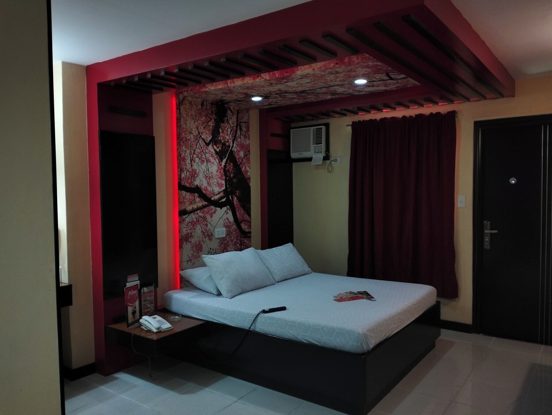 A bedroom with a bed, a nightstand with a phone, a wall-mounted air conditioner, and a door.