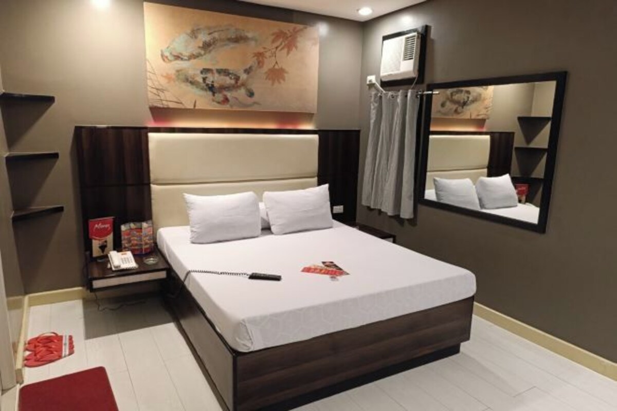 A modern hotel room with a large bed, a bedside table with a phone and magazines, a wall-mounted air conditioner, and a large mirror.