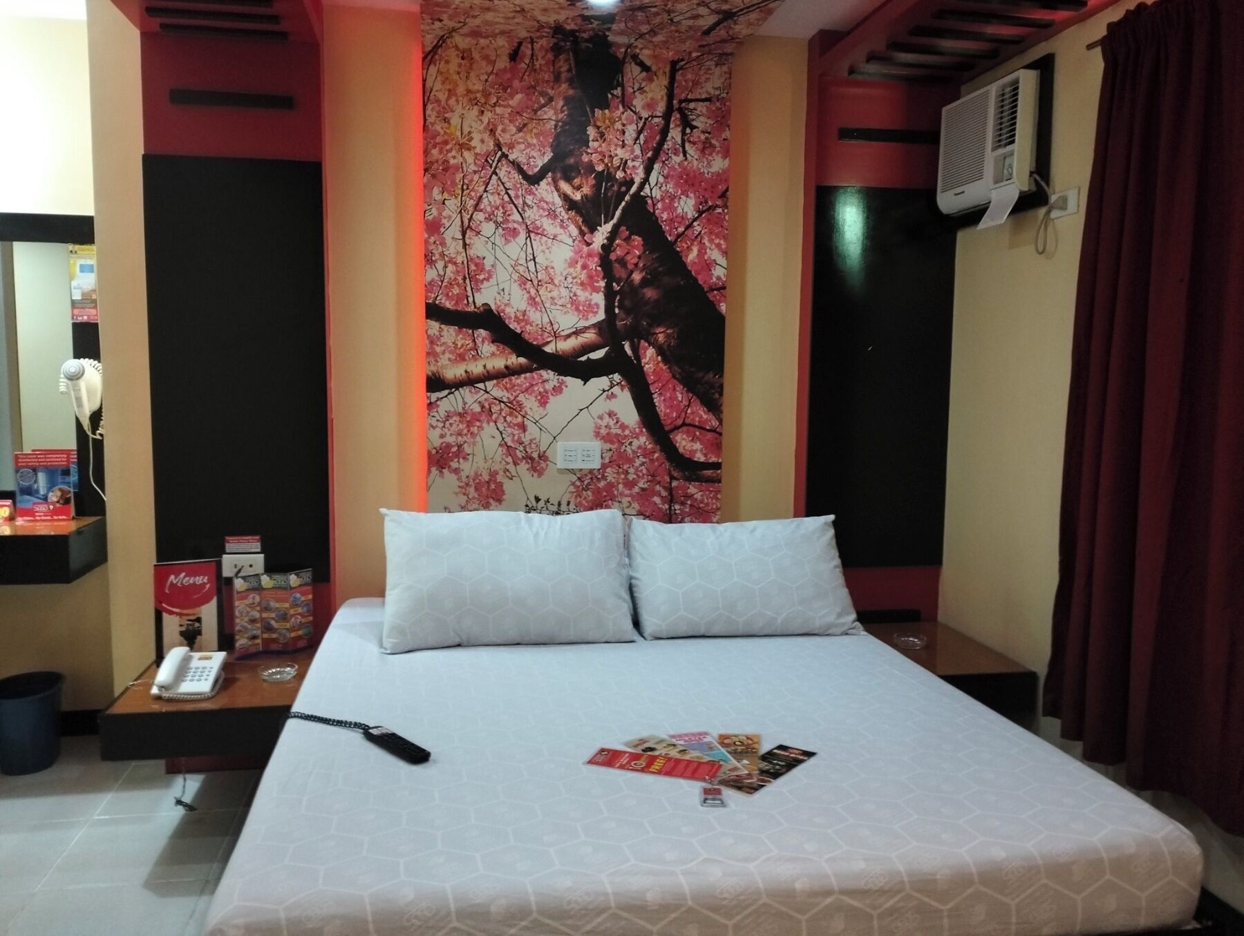 A hotel room with a bed, a wall-mounted air conditioner, a telephone, and a magazine on the bed.