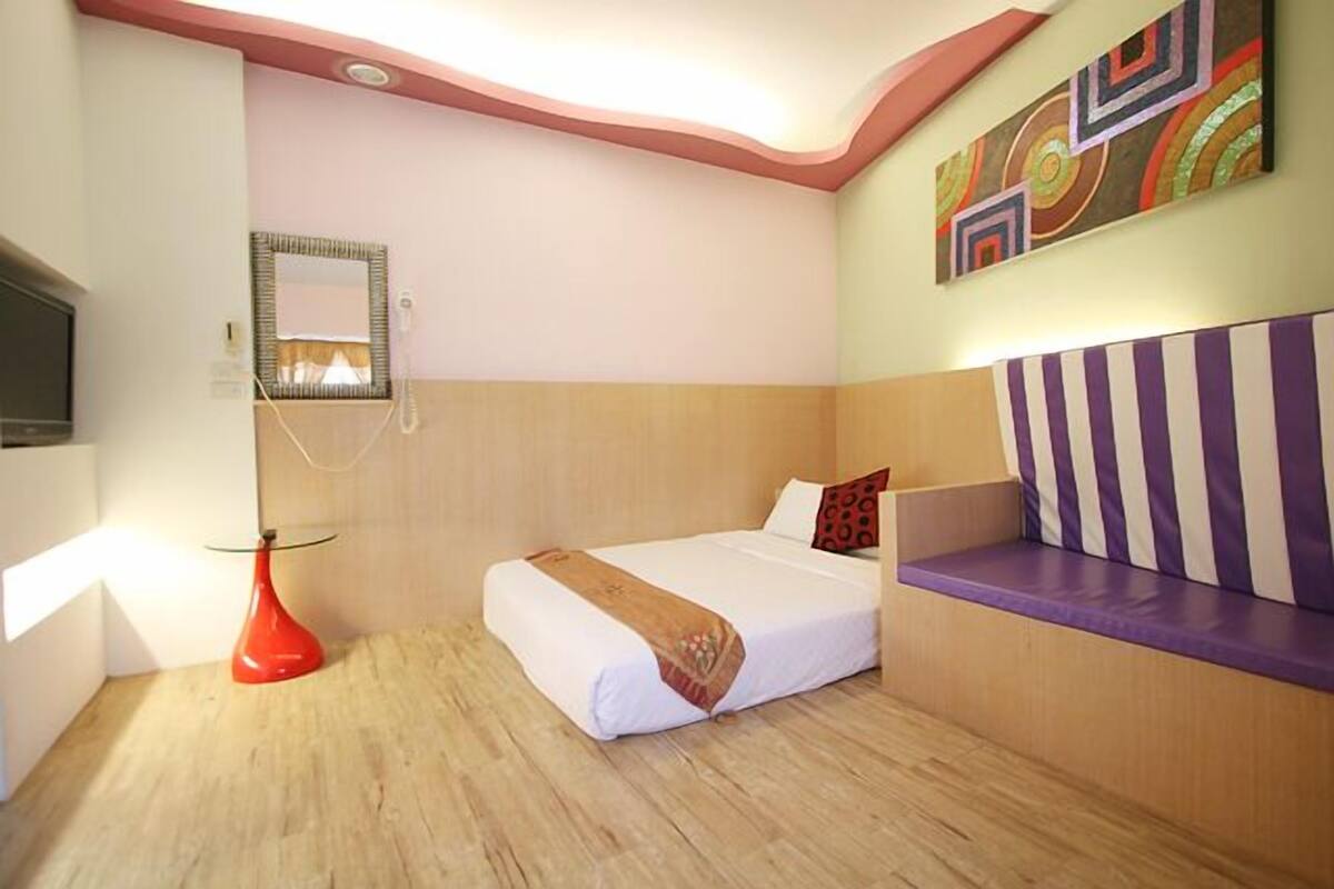 A hotel room with a bed, a red vase, a television, and a wall with colorful artwork.