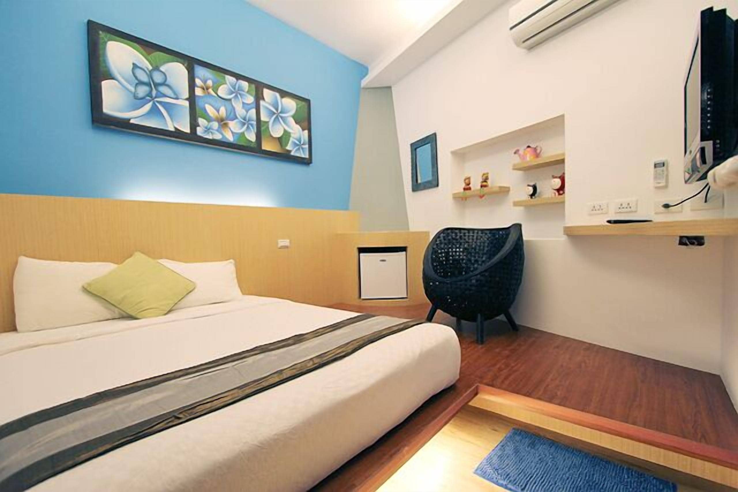 A hotel room with a bed, a chair, a desk, and a shelf with decorative items.