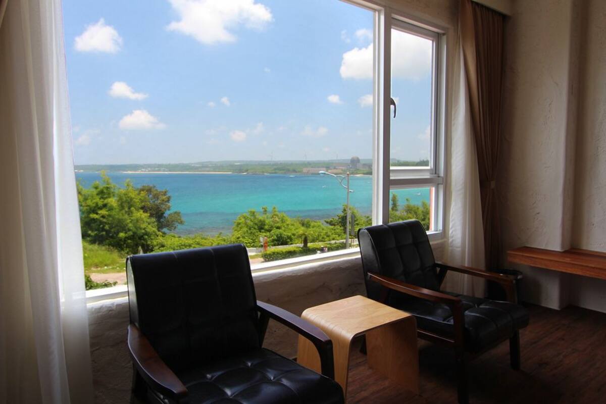 A room with two leather chairs, a wooden table, and a large window offering a view of the sea and greenery.