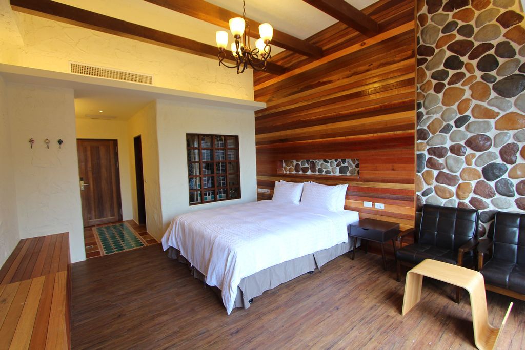A bedroom with a large bed, wooden flooring, a stone wall, and a seating area with chairs.