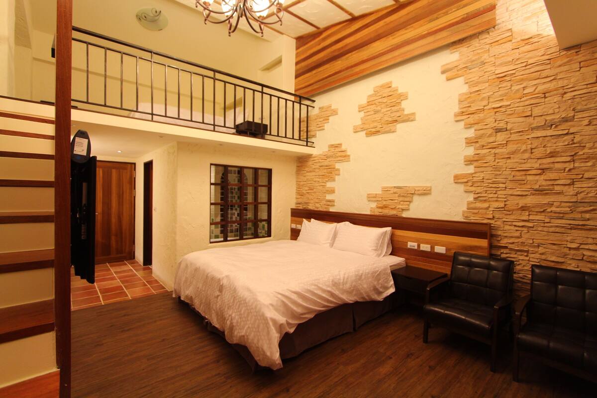 A bedroom with a large bed, a wooden headboard, two black leather chairs, a stone wall, and a balcony with a railing.