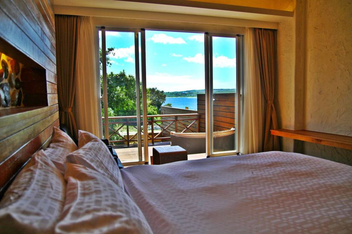 A bedroom with a large bed, a wooden headboard, and a view of a balcony with a table and chairs.