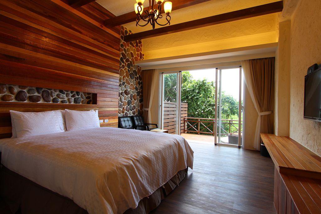 A bedroom with a large bed, stone wall, wooden ceiling, and a balcony with a view.