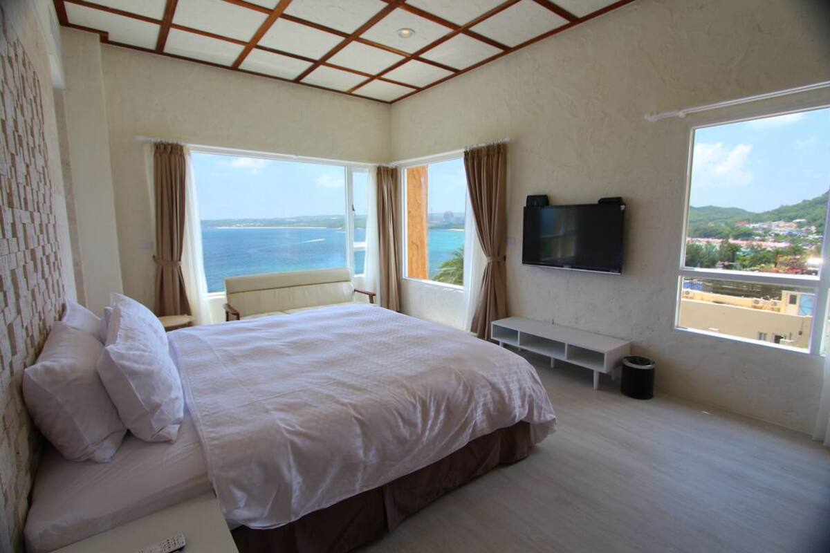 A bedroom with a large bed, a TV, a bench, and a view of the sea.