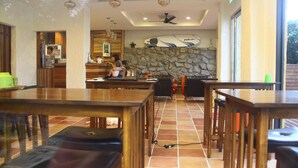 Un restaurante con mesas y sillas de madera, una pared de piedra decorada con una tabla de surf, y una zona de cocina visible a través de un vano abierto.