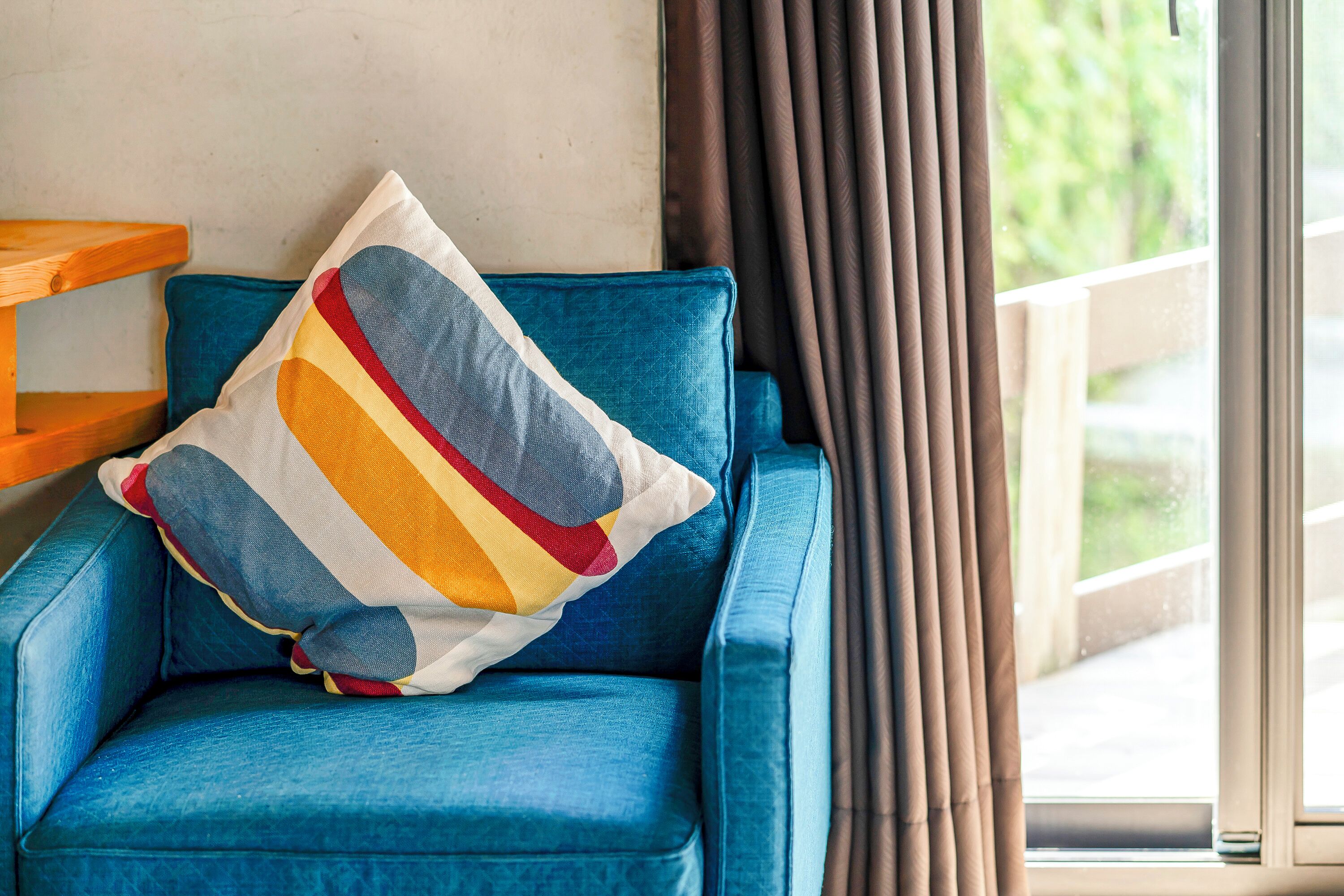 A blue sofa with a colorful pillow, a wooden side table, and a view of greenery through a window with curtains.