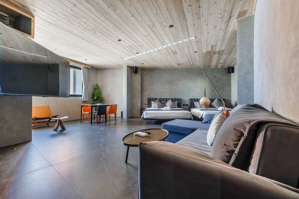 A modern hotel room with a wooden ceiling, a large bed, a sofa, a small table, and a dining area with chairs.