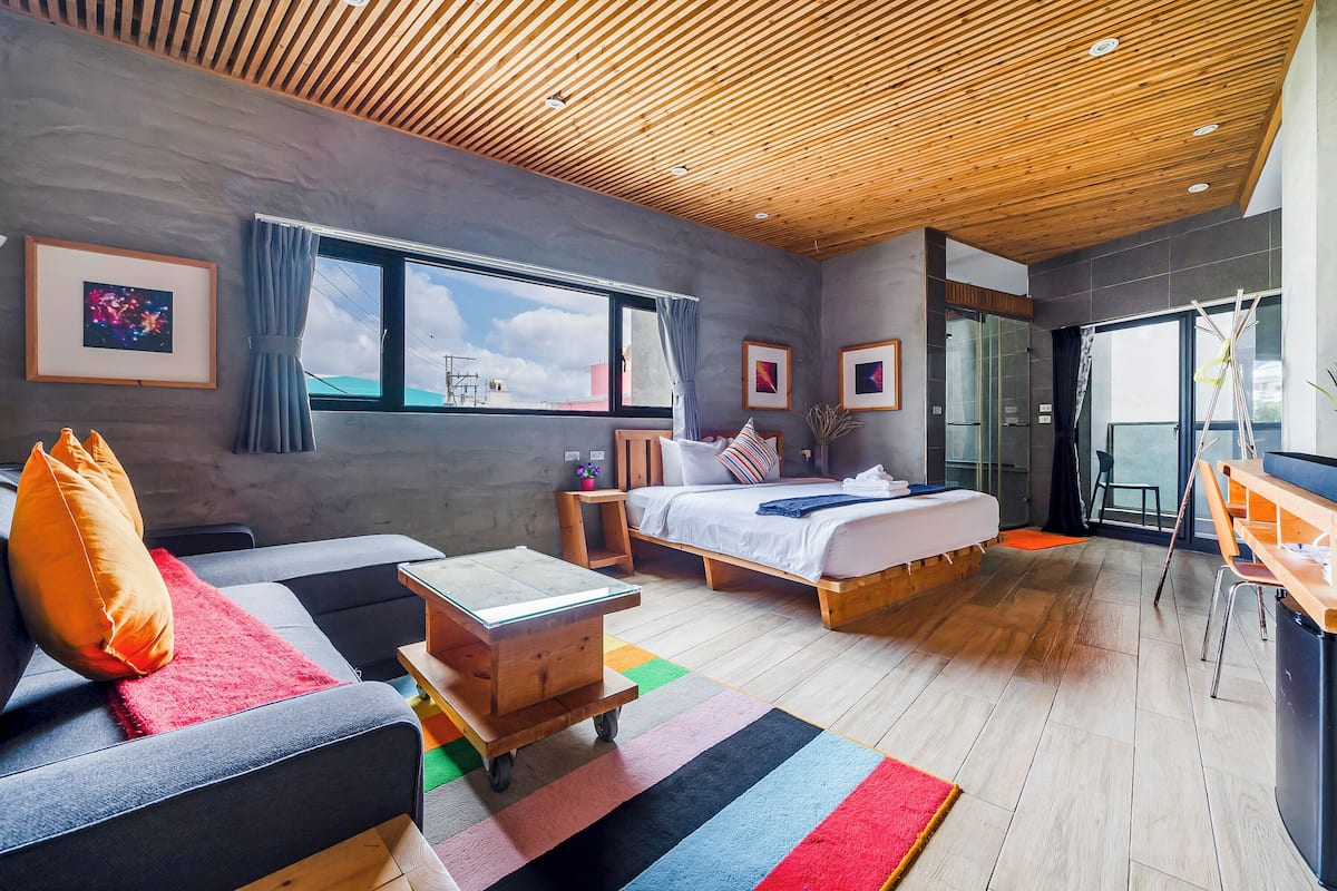 A modern hotel room with a large bed, a sofa, a small table, and a desk. There are multiple windows and a view of the outside.