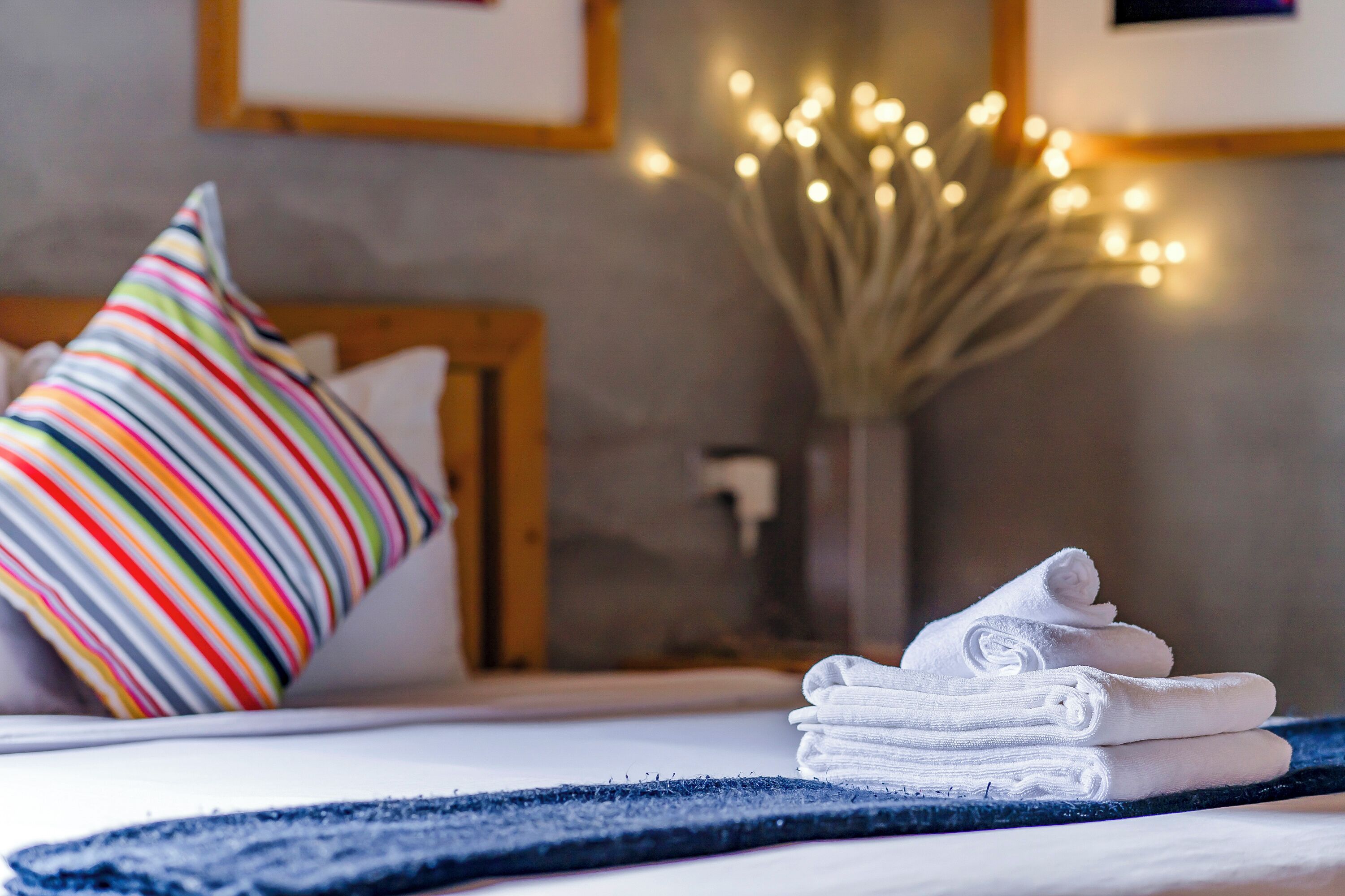 A neatly made bed with white towels, a colorful striped pillow, and a vase with lights in the background.