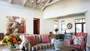 A spacious living room with a sofa, armchair, and coffee table. There is a large painting on the wall and a vase with flowers on a side table. - Kingsmead Guest House (Harare)