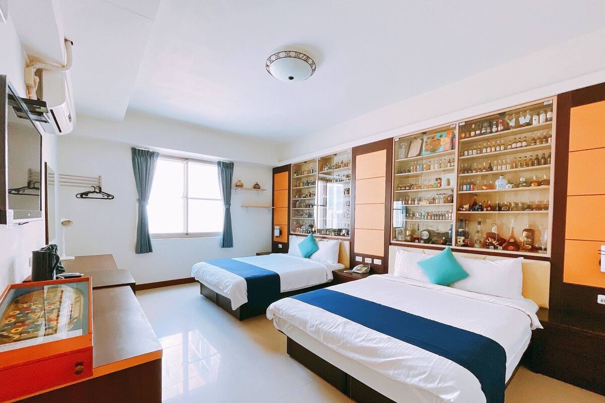 Ein Hotelzimmer mit zwei Betten, einer Minibar, einem großen Fenster mit Vorhängen und einem an der Wand befestigten Regal mit Flaschen.