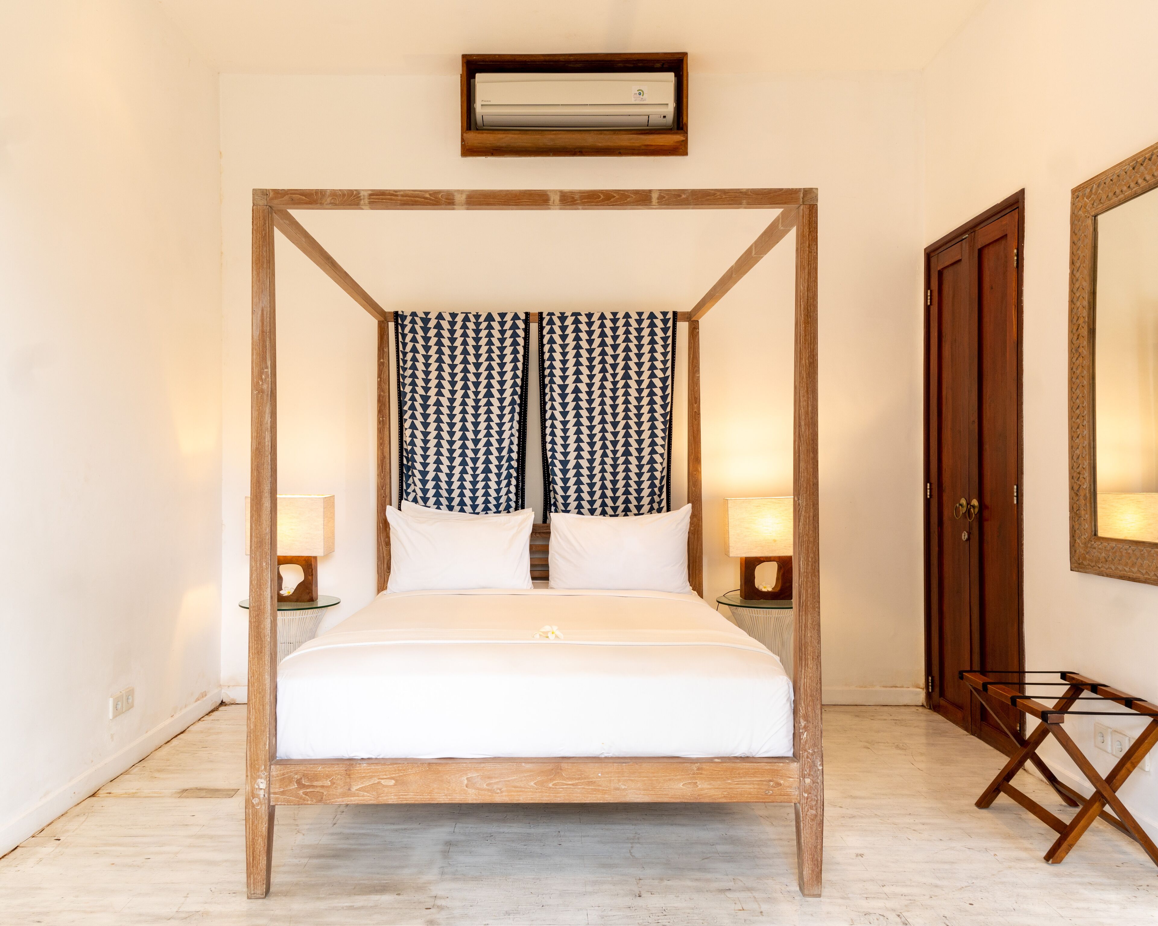 A four-poster bed with white bedding, two bedside lamps, patterned curtains, a wooden floor, a wall-mounted air conditioner, and a wooden door.