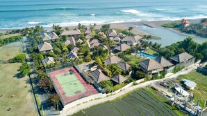 Eine Vogelperspektive zeigt einen Küstenort mit mehreren bungalowartigen Gebäuden, einem Tennisplatz und einem Strand.
