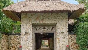 Ein steinernes Portal mit einem Strohdach, das von Statuen gesäumt und von Grünpflanzen umgeben ist.