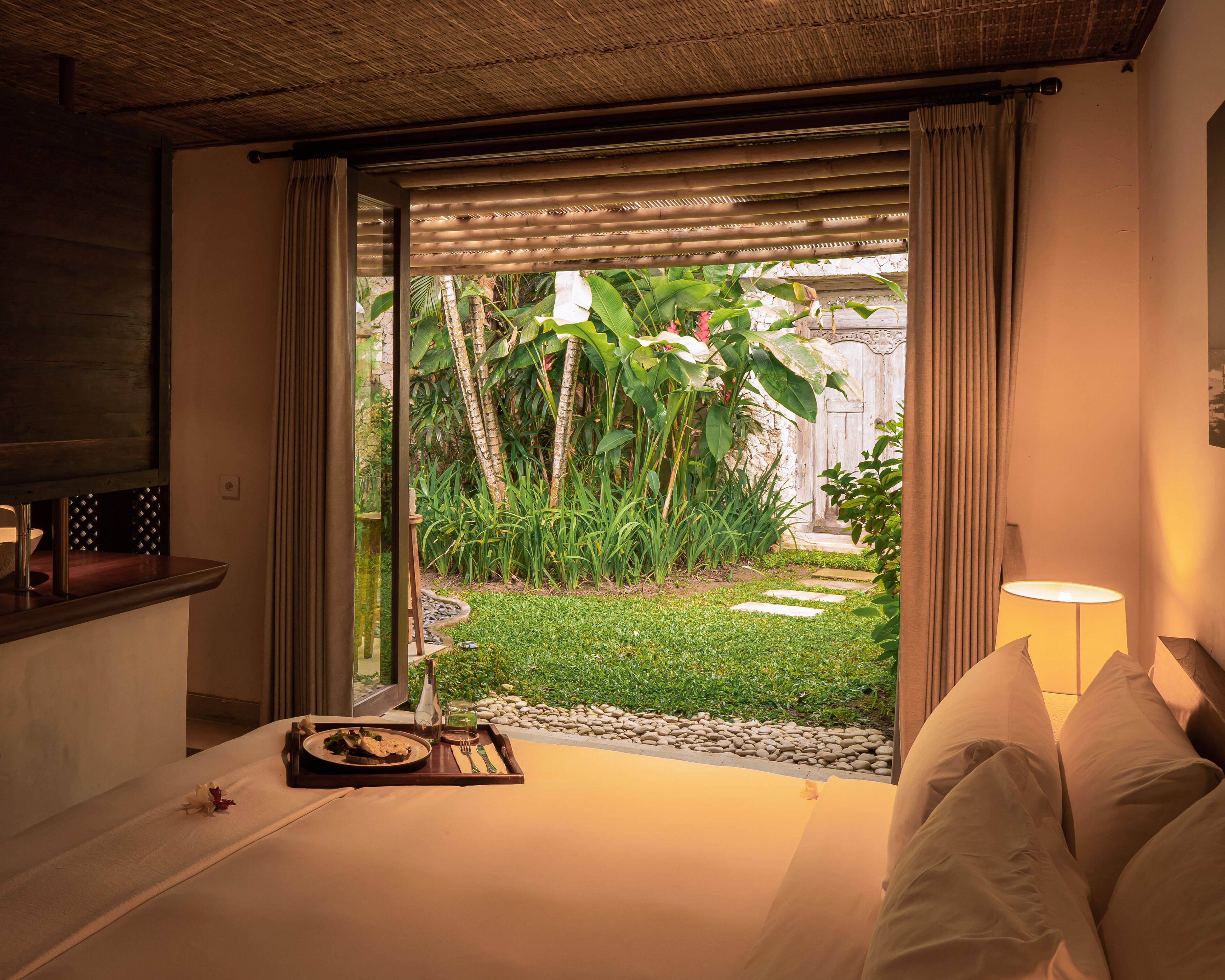 A hotel room with a bed, a bedside table with a lamp, and a view of a garden with plants and a pathway.
