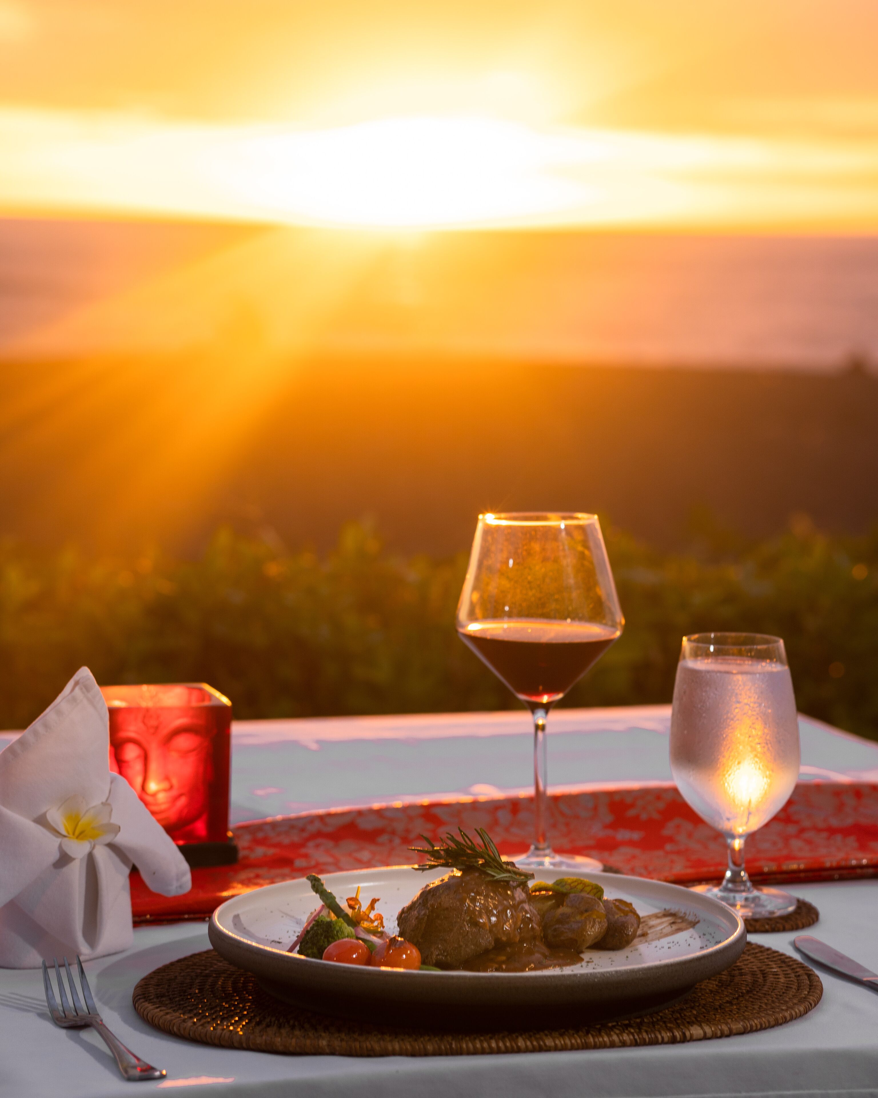 Une table dressée pour un repas, avec une bougie, un verre de vin et un verre d’eau, et en arrière-plan, un magnifique coucher de soleil.