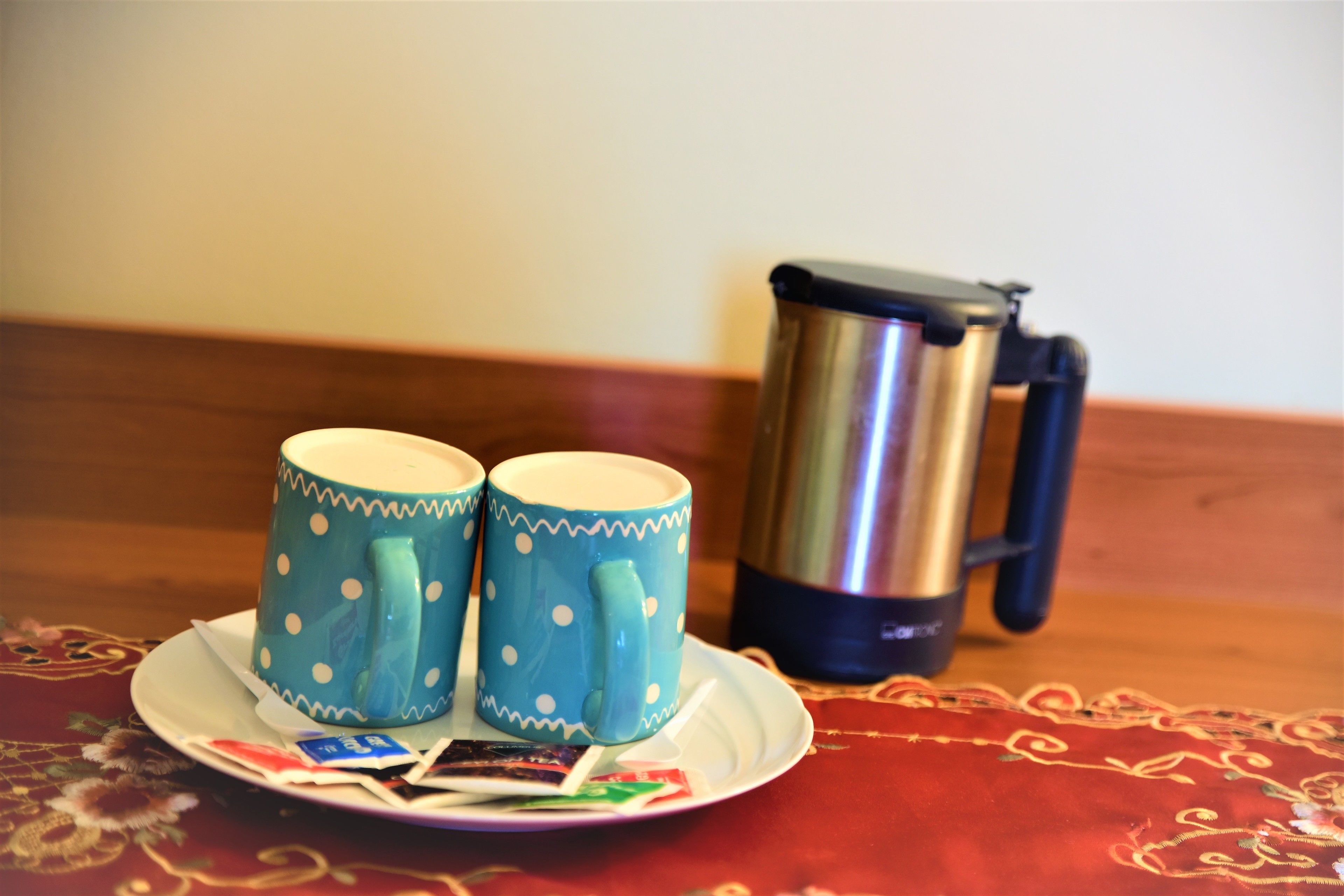 Two polka-dotted mugs on a plate with a thermos and some packets on a table with a patterned tablecloth.