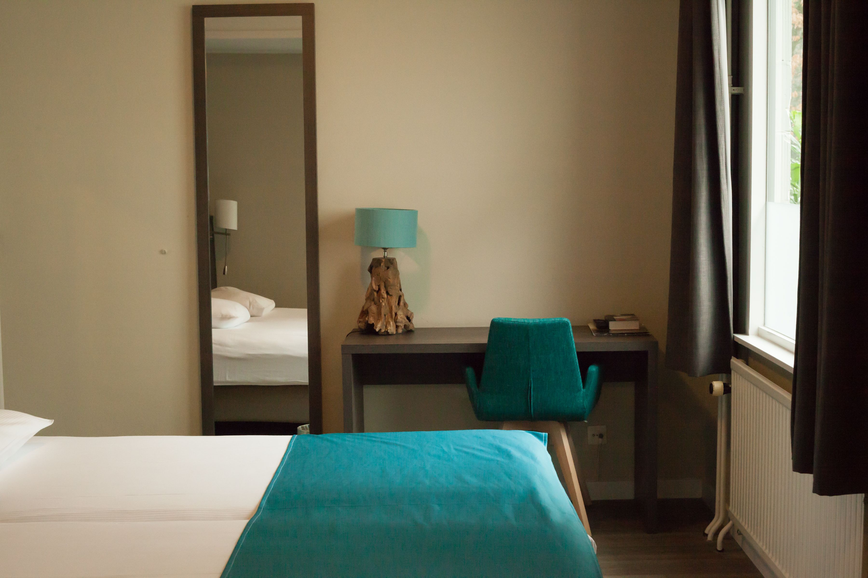 Una habitación de hotel con cama, escritorio, silla, lámpara y ventana con cortinas.