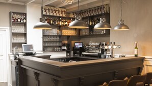 Zona de bar con una barra central, taburetes, estantes con botellas y vasos, e iluminación colgante.