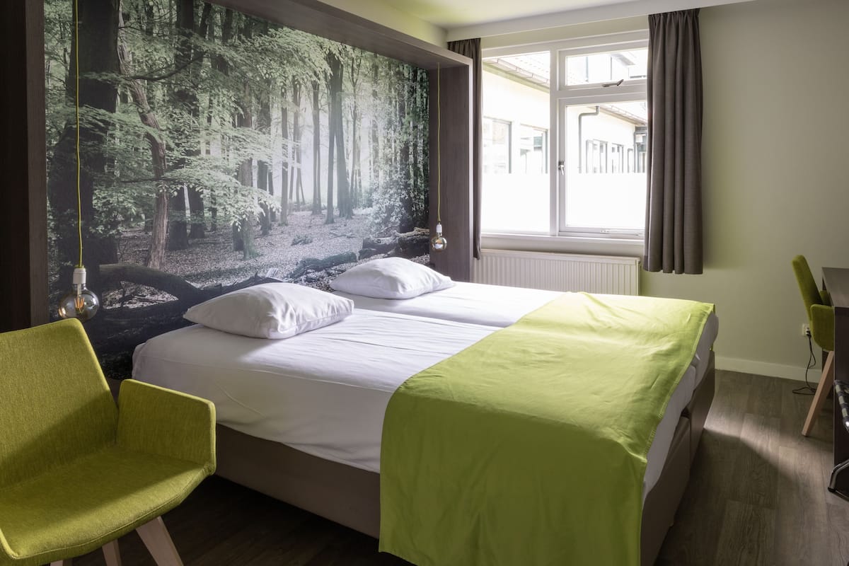 Habitación de hotel con cama, dos sillones verdes, un mural grande con un bosque, una ventana con cortinas y un escritorio con silla.