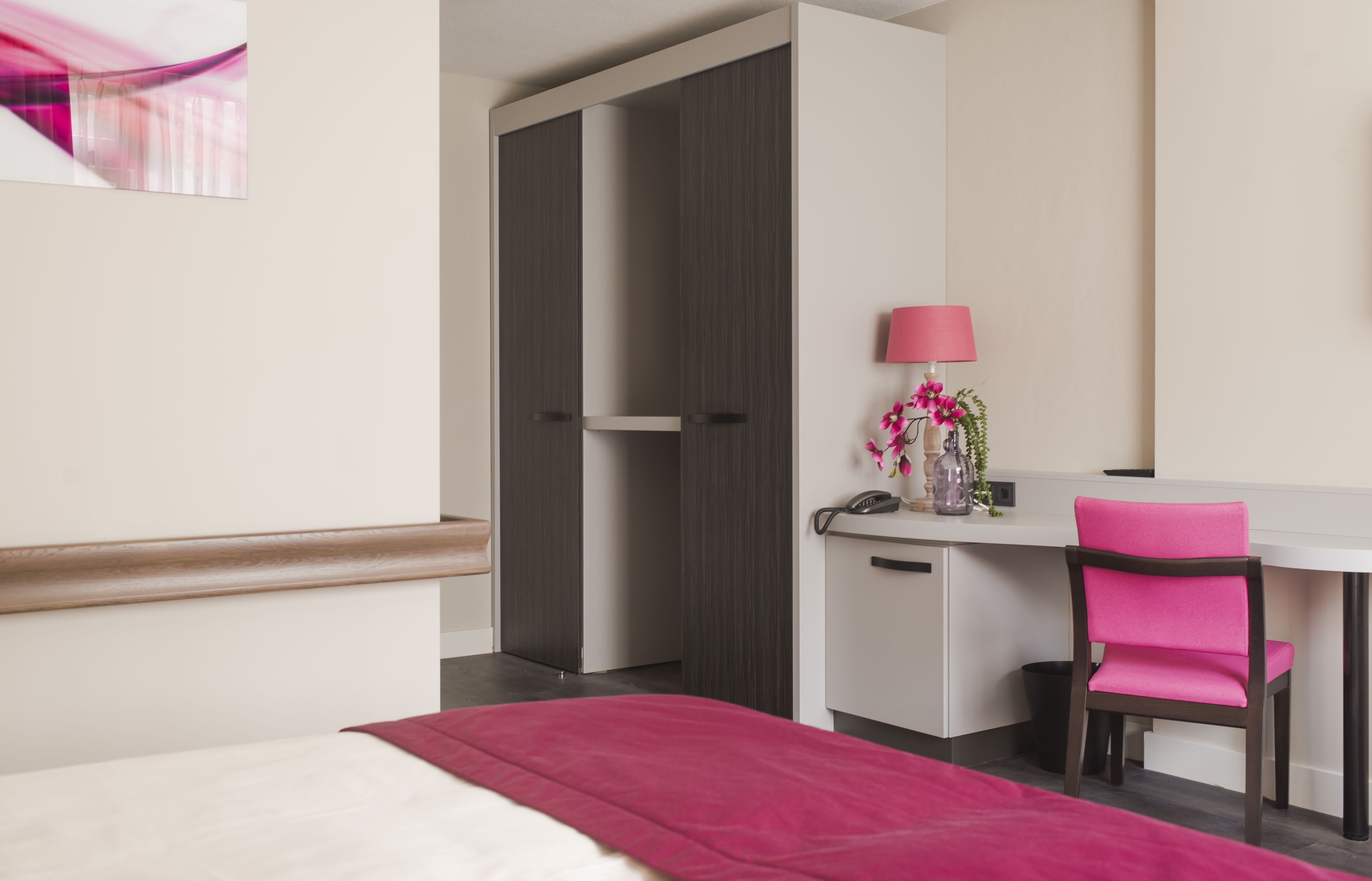 Una habitación de hotel con cama, escritorio, silla, lámpara y una planta pequeña.