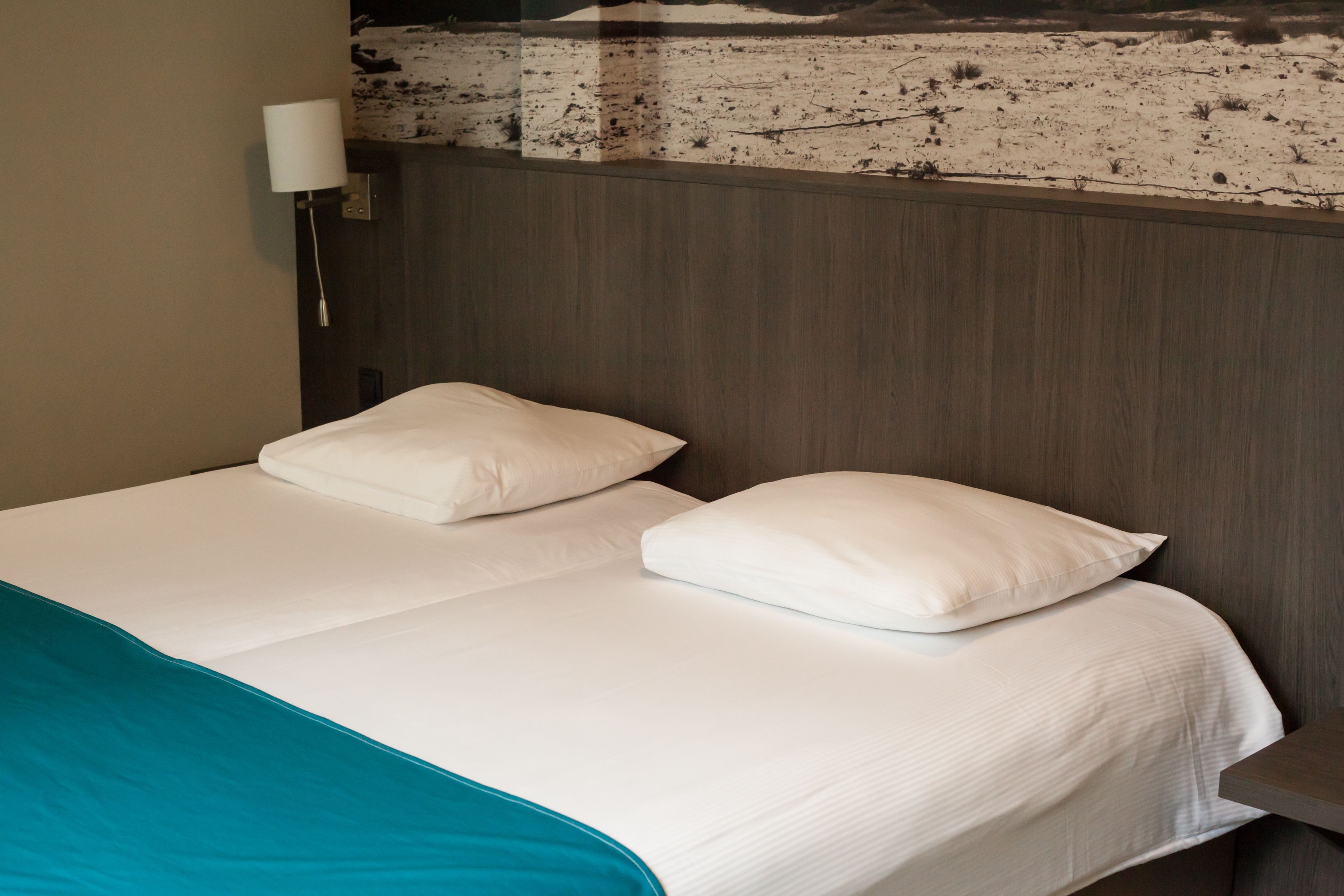 A hotel room with a bed, two pillows, a teal throw blanket, a wall-mounted lamp, and a textured wall above the bed.