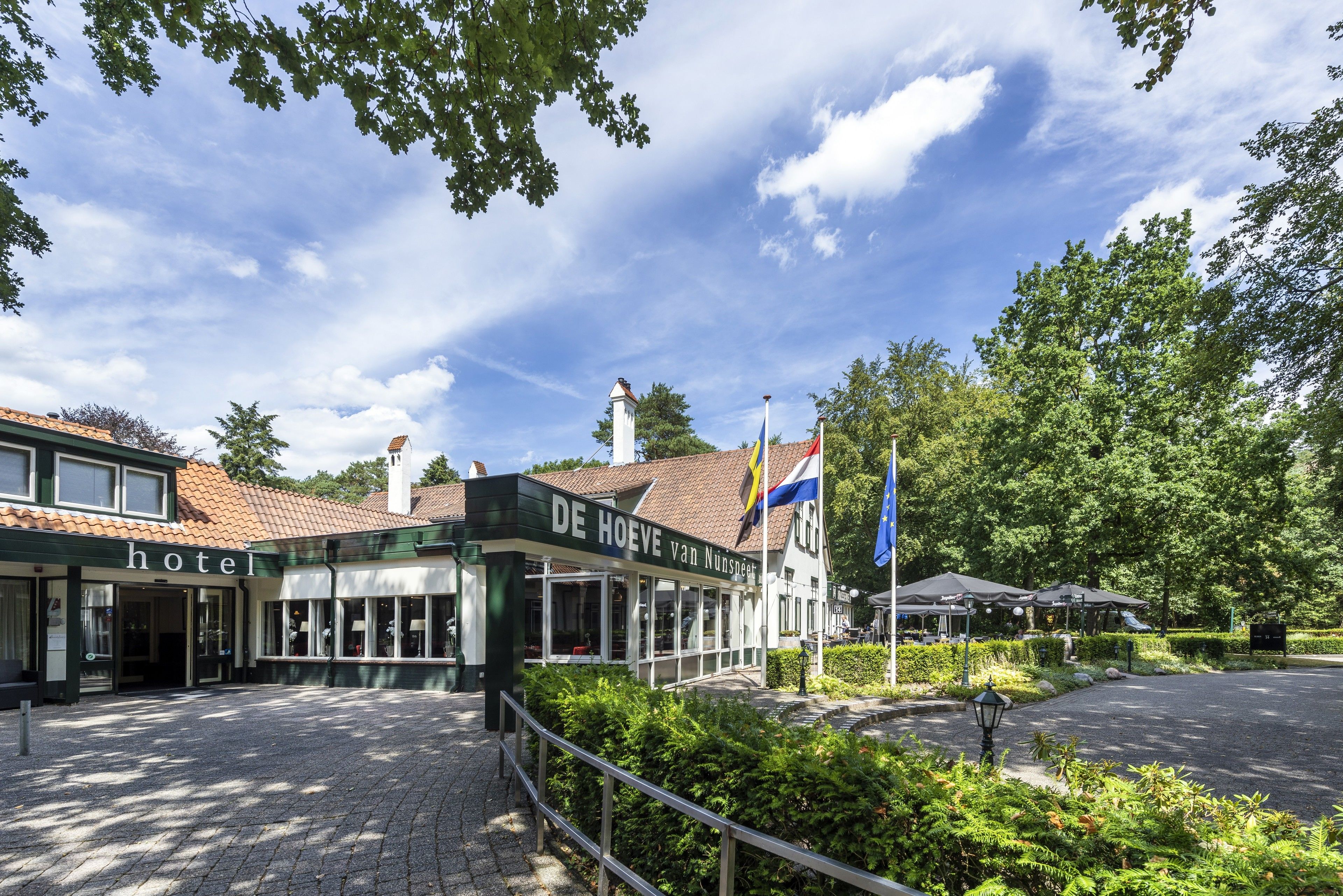Een hotel met een pannendak, een bord waarop staat 'DE HOEVE van Nunspeet' en vlaggen.