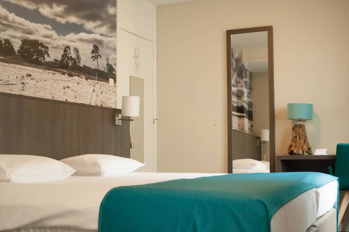 Habitación de hotel con cama, escritorio, espejo y vista a la playa desde la ventana.