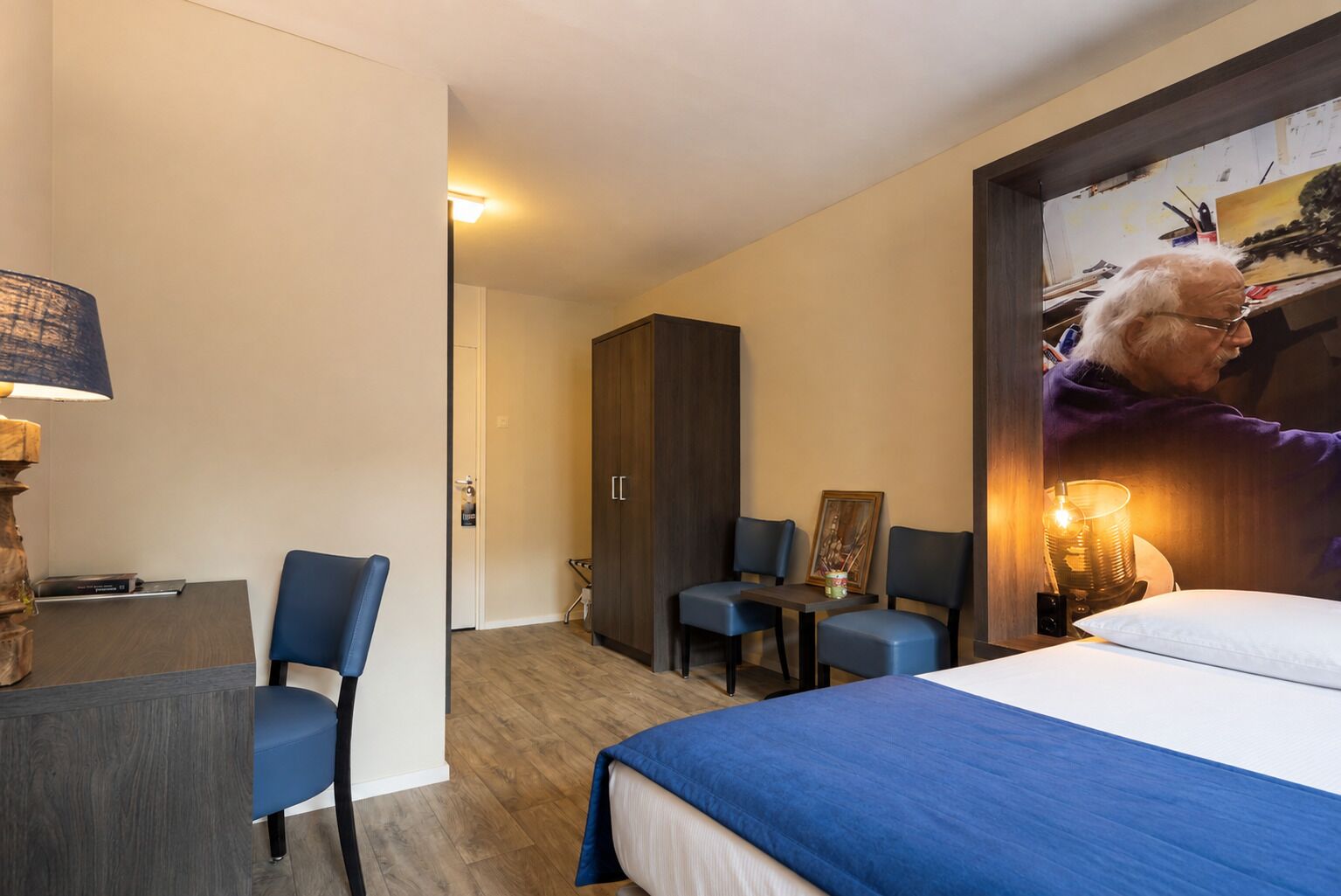Ein Hotelzimmer mit Bett, Schreibtisch, zwei Stühlen, einem Kleiderschrank und einem Bild an der Wand.